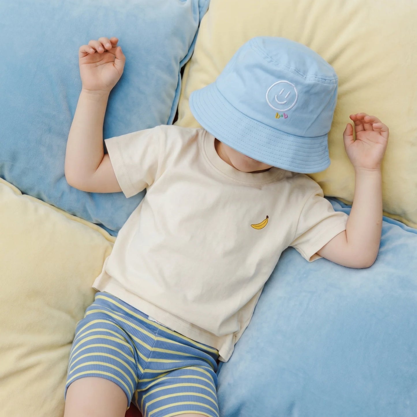 Child wearing a light blue bucket hat and striped shorts lying on cushions.
