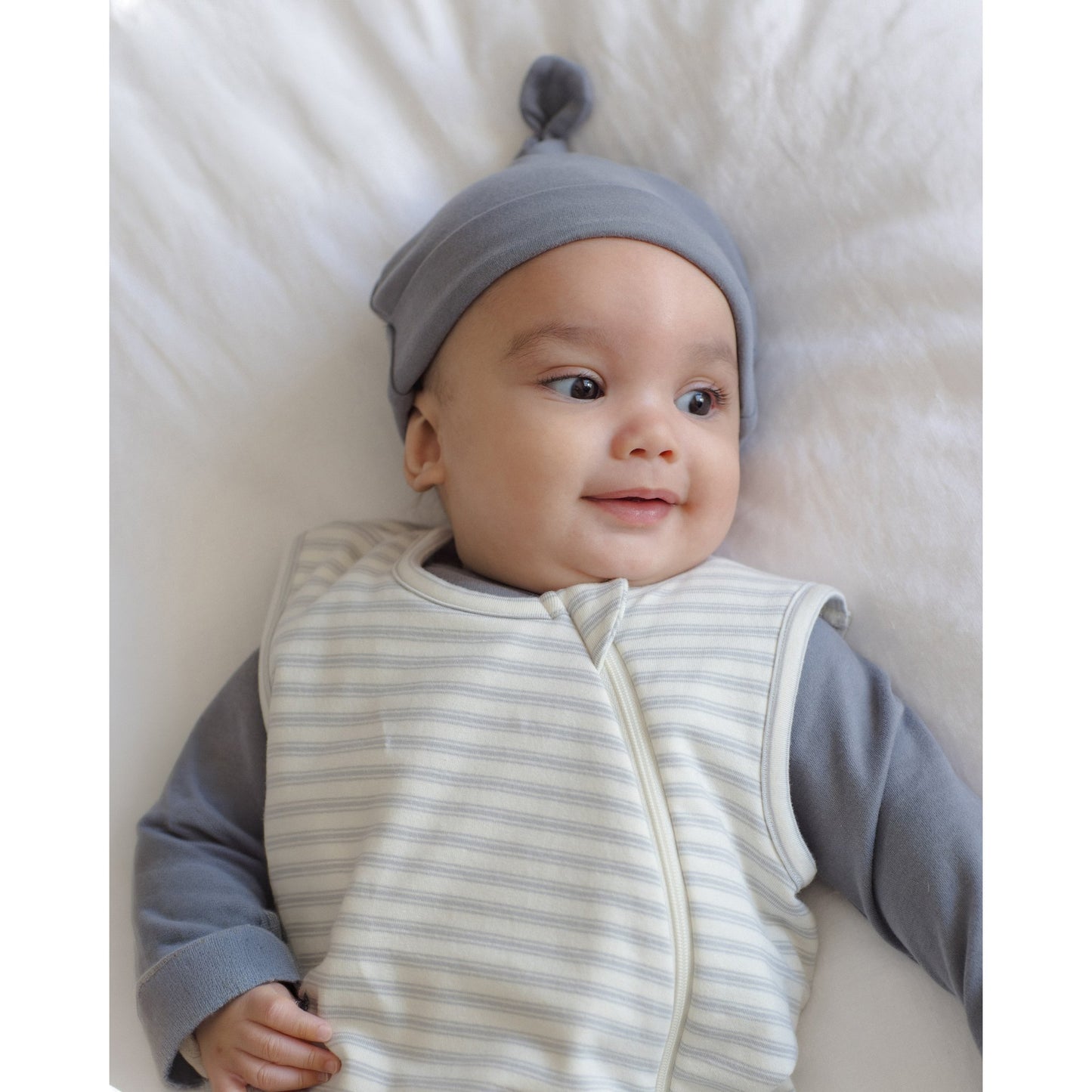 A baby lying on a white textured blanket, wearing a gray knit pom-pom bonnet and a sleeveless zip-front sleep vest with thin gray stripes over a gray long-sleeve shirt.