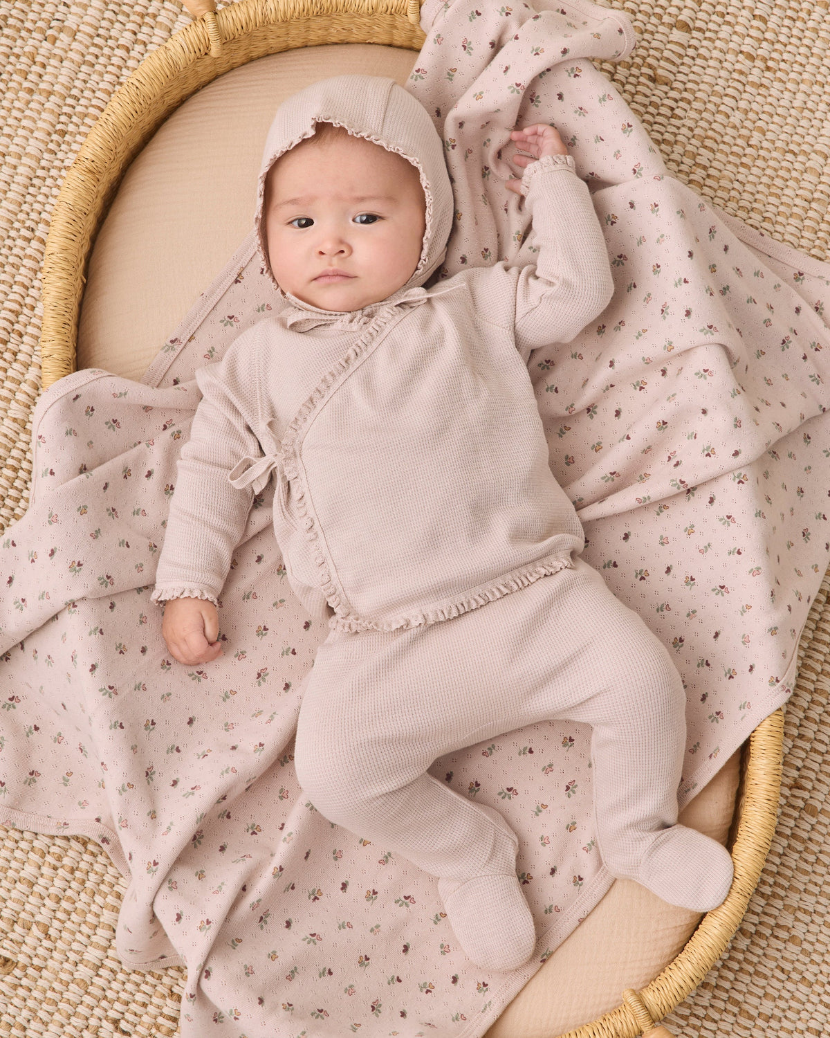 A baby wearing a light beige footed onesie and bonnet lies on a cream woven mat, wrapped in a matching beige and pink floral blanket.