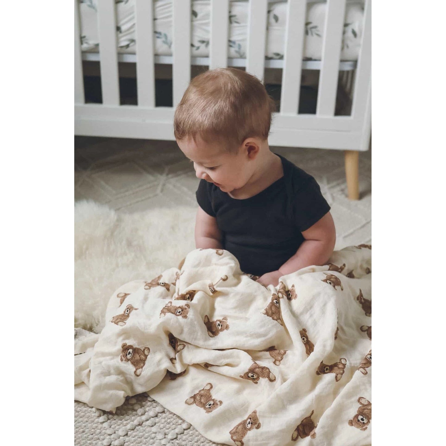 Child sitting on a rug with a white blanket featuring brown bear patterns, in front of a white crib.