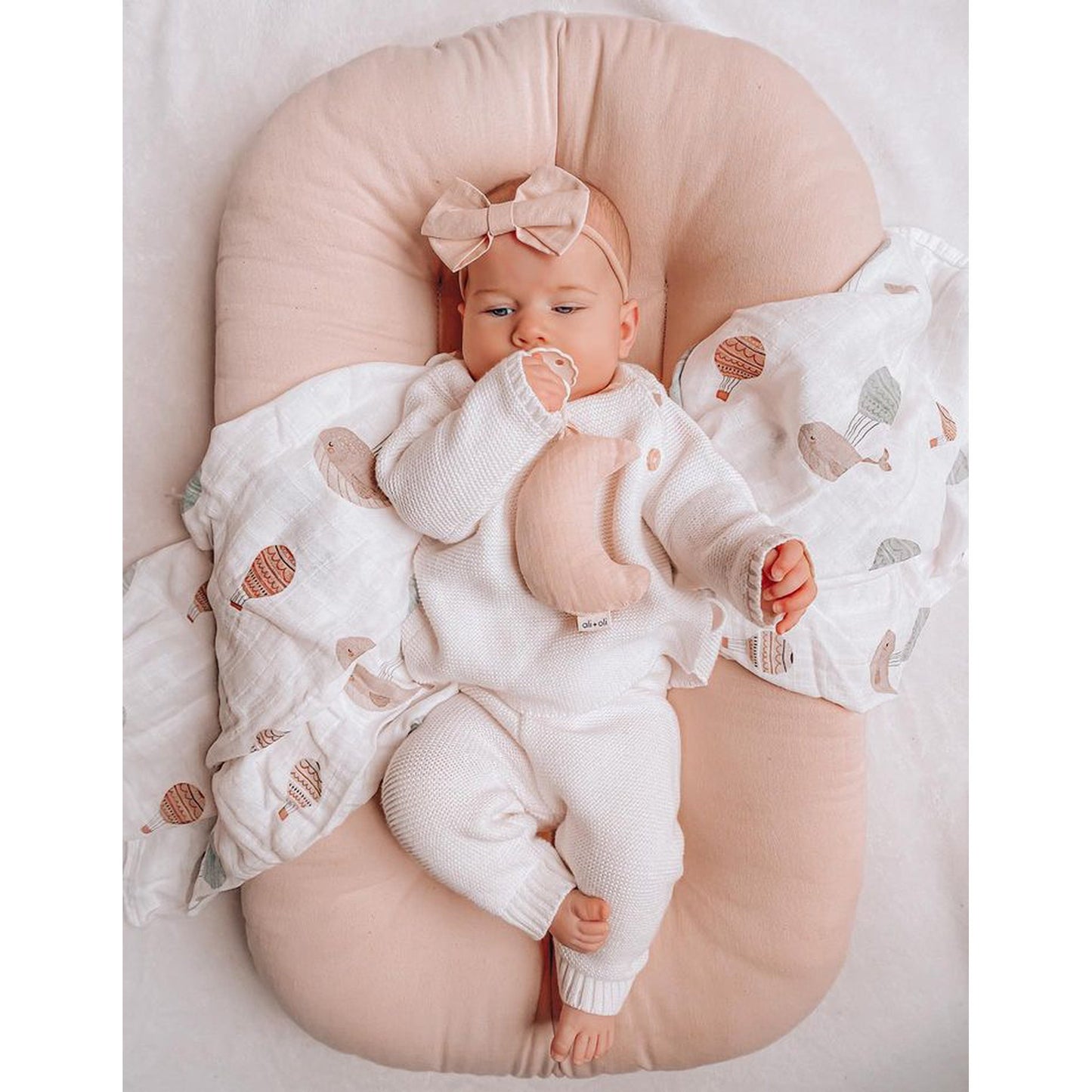 Baby lying on a pink pillow with a pacifier and bow headband, wrapped in a white blanket with animal patterns.