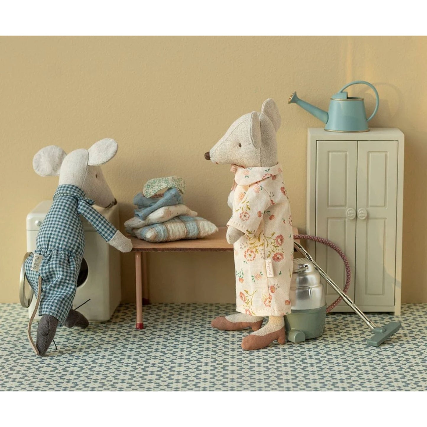 Two toy mice in a miniature room setting with a washing machine and cabinet.