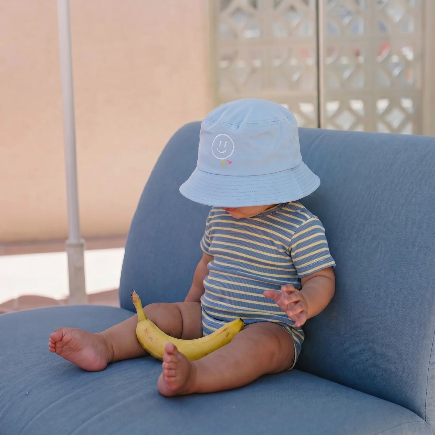 Baby sitting on a blue chair wearing a blue hat and striped shirt, holding a banana.