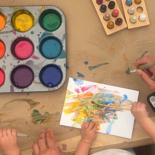A top-down view of a painting activity on a wooden table. A child's hands and an adult's hands are visible, painting on white paper.