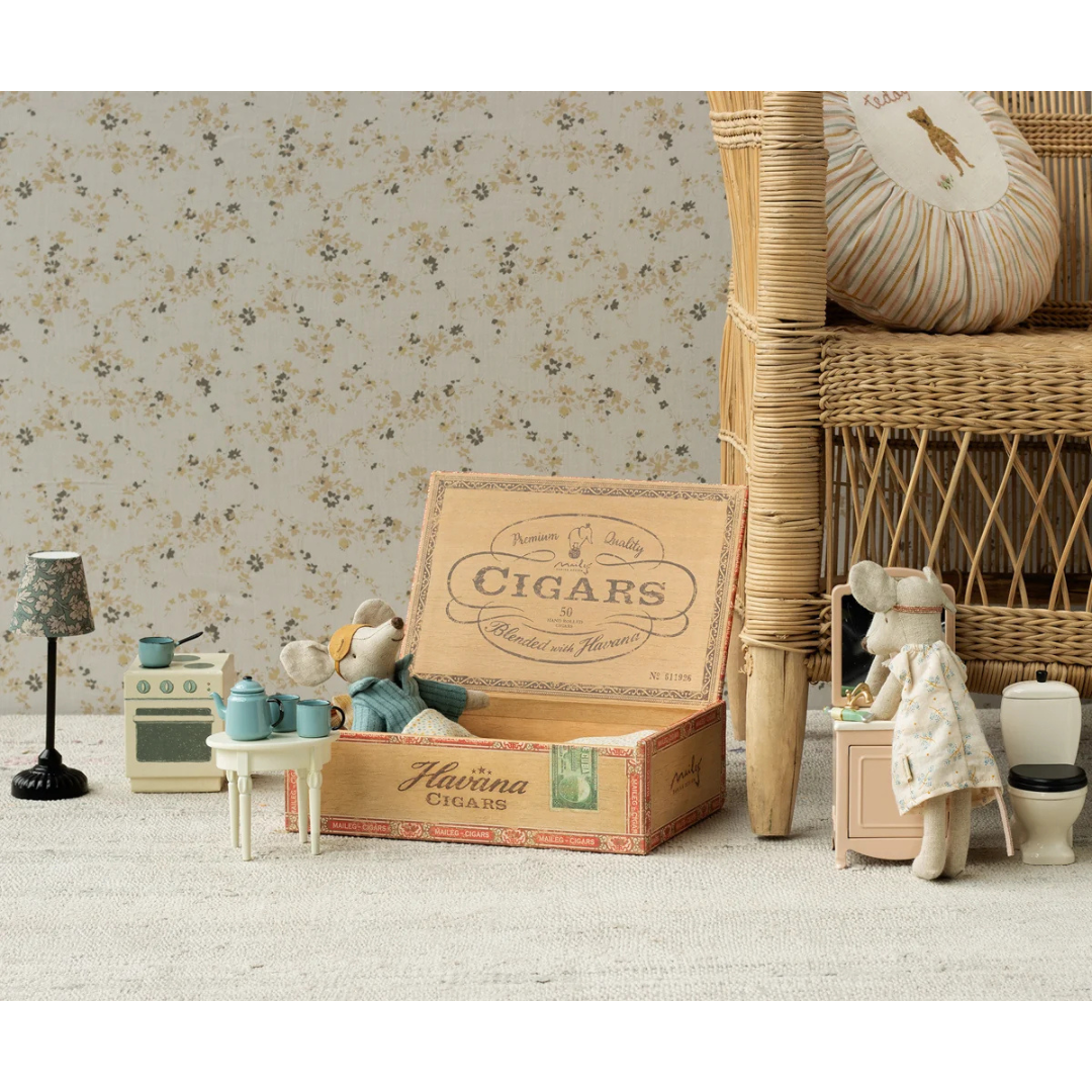 A vintage-style toy or collectible cigar box labeled 'Macrina Cigars' sits open on a light rug, styled in a dollhouse-like interior with miniature furniture, a teddy bear, and a doll. A wicker chair and floral wallpaper are visible in the background.