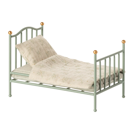 A miniature metal daybed with a sage green frame and gold ball finials, styled with a textured cream faux fur throw and matching pillow on a white background.