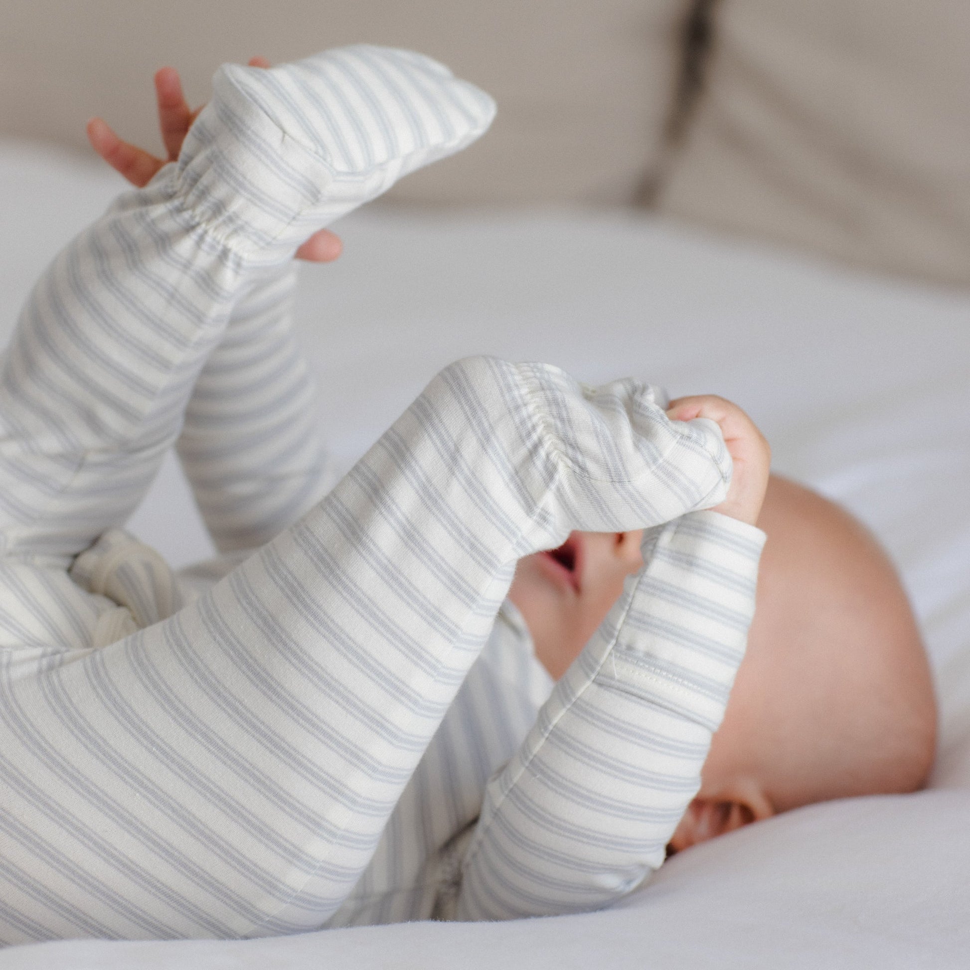 A baby lying on a white bed, wearing a long-sleeve footed sleeper with thin gray and white stripes.