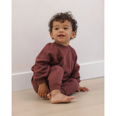 A toddler with curly dark hair sits barefoot on a light wood floor against a white wall, wearing a matching rust-brown sweatshirt and jogger pants set.
