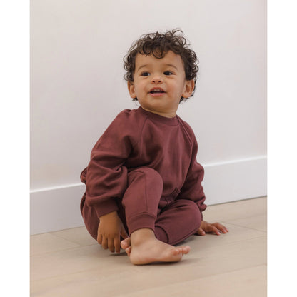 A toddler with curly dark hair sits barefoot on a light wood floor against a white wall, wearing a matching rust-brown sweatshirt and jogger pants set.