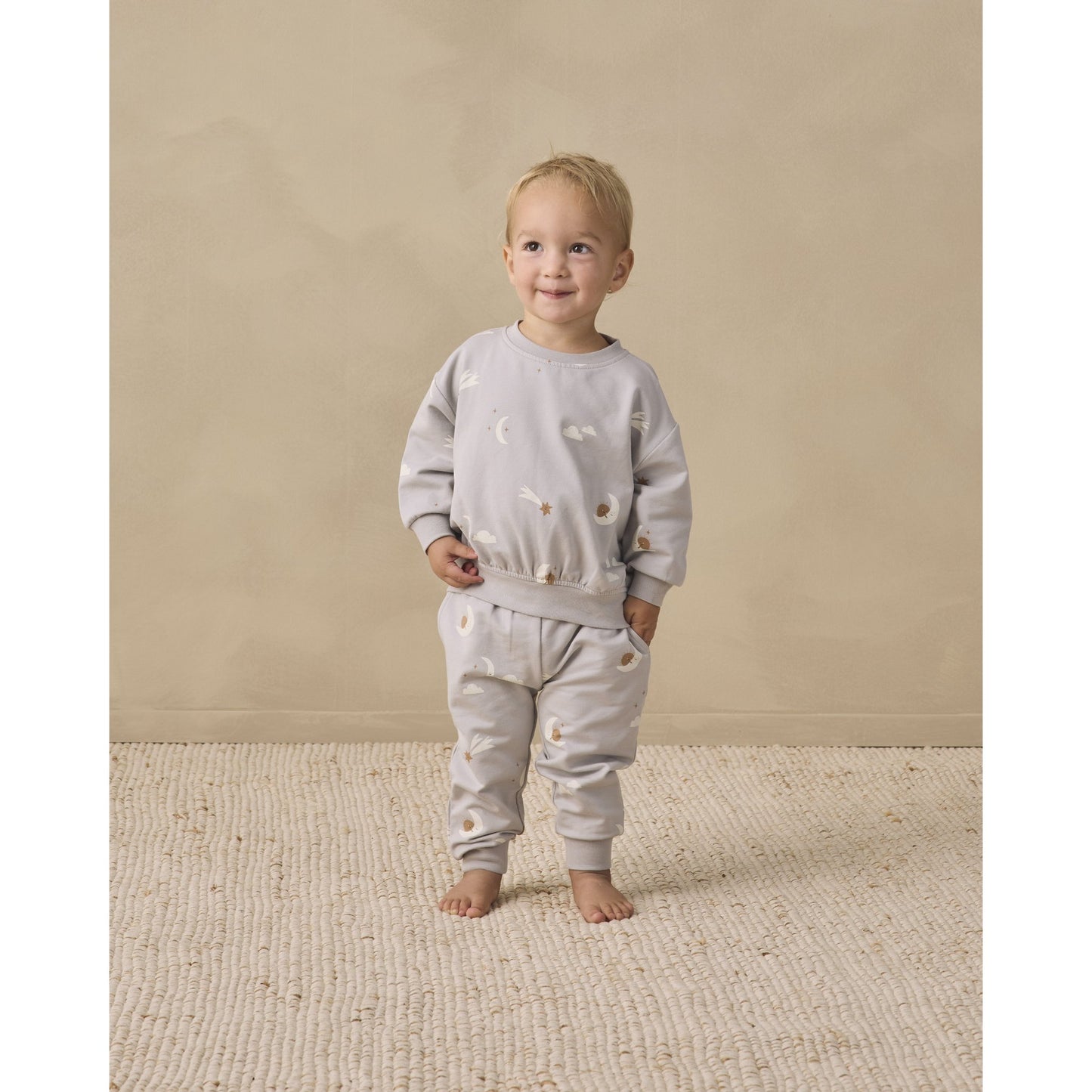 A toddler stands barefoot on a textured cream rug against a warm beige studio wall, wearing a light gray two-piece outfit with sky friends print.