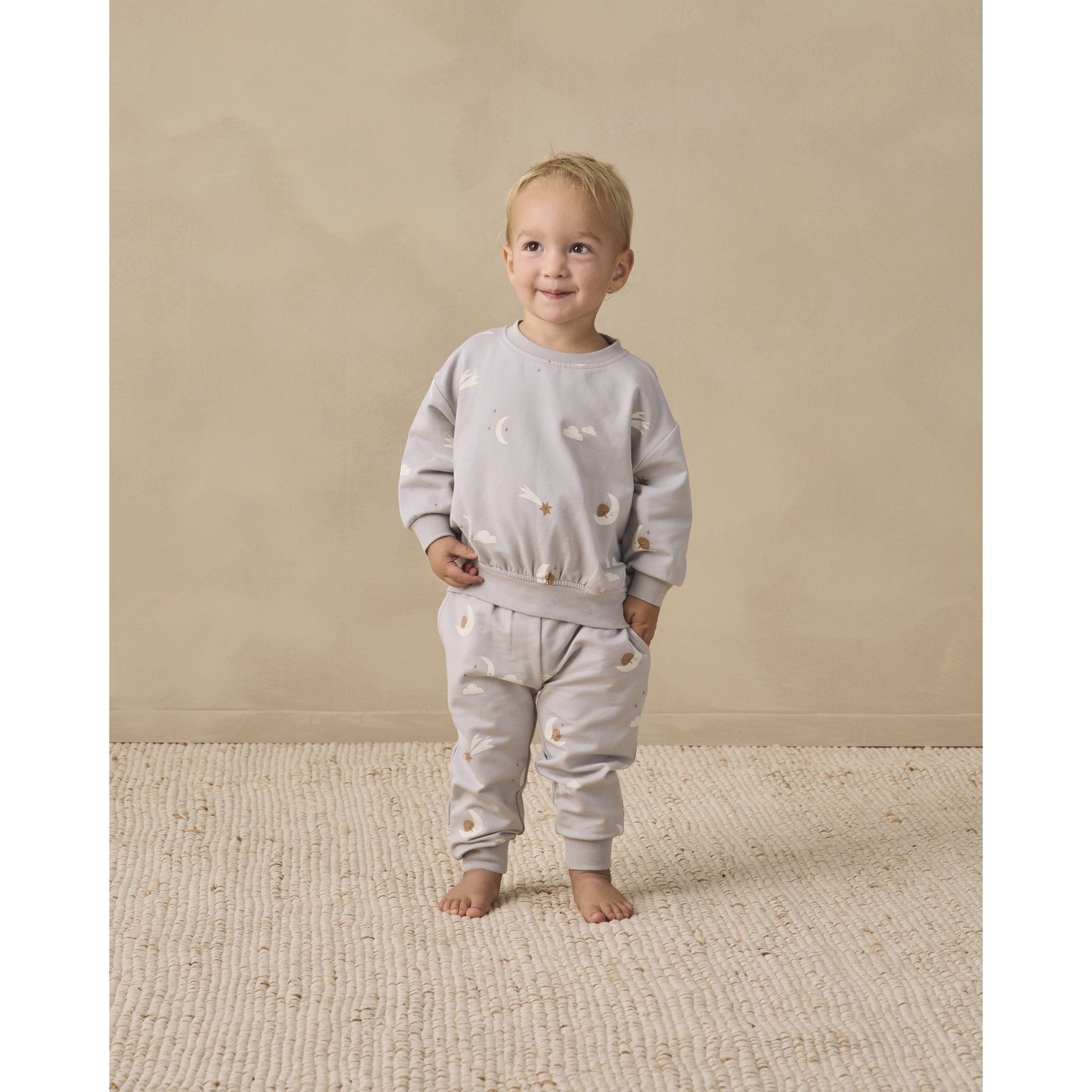 A toddler stands barefoot on a textured cream rug against a warm beige studio wall, wearing a light gray two-piece outfit with sky friends print.