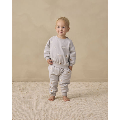 A toddler stands barefoot on a textured cream rug against a warm beige studio wall, wearing a light gray two-piece outfit with sky friends print.