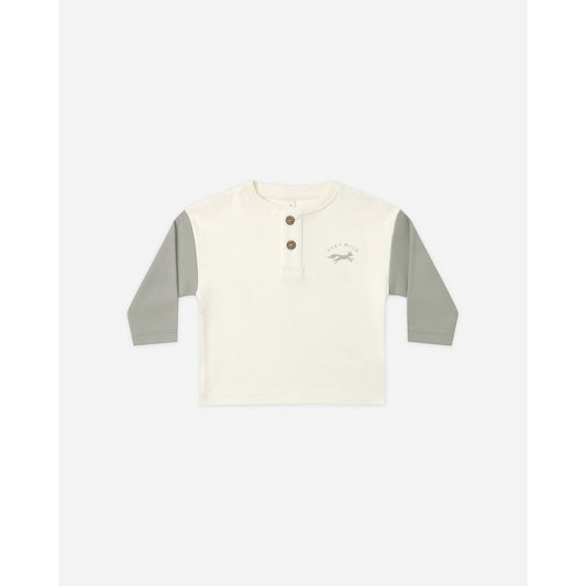A toddler's long-sleeve henley shirt in off-white with sage green sleeves, featuring a three-button placket and a small graphic on the left chest.