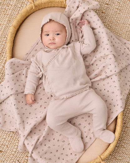 A baby wearing a light beige footed onesie and bonnet lies on a cream woven mat, wrapped in a matching beige and pink floral blanket.