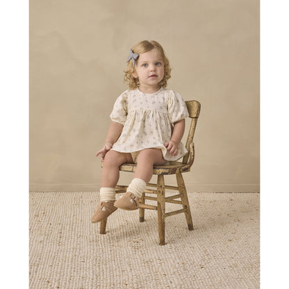 A toddler girl with blonde curly hair wearing a cream short-sleeve dress with a tiny floral print, sitting on a rustic wooden chair against a neutral beige background.