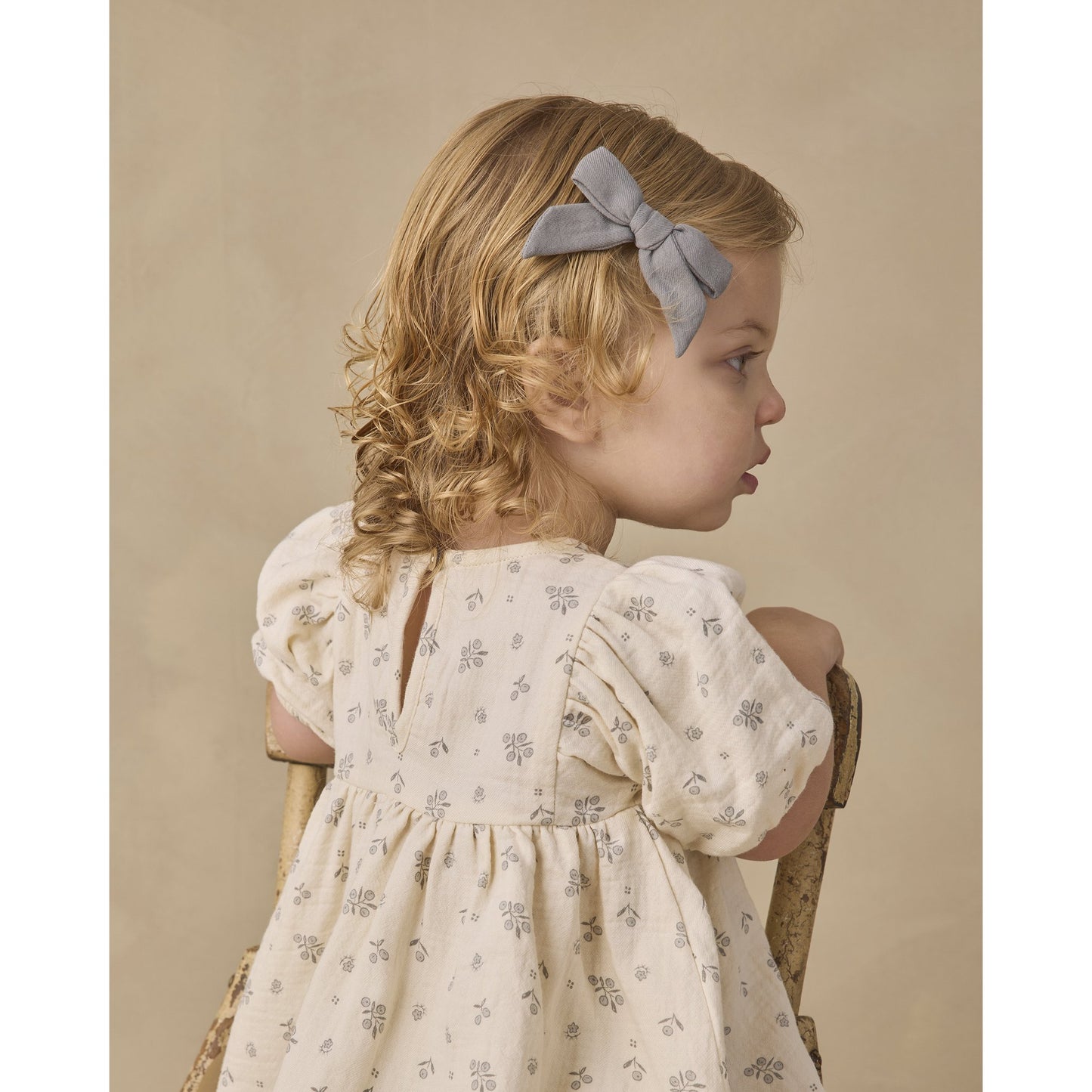 A young girl with blonde curly hair wearing a light gray bow headband and a cream-colored floral dress, sitting on a wooden chair against a warm beige background.