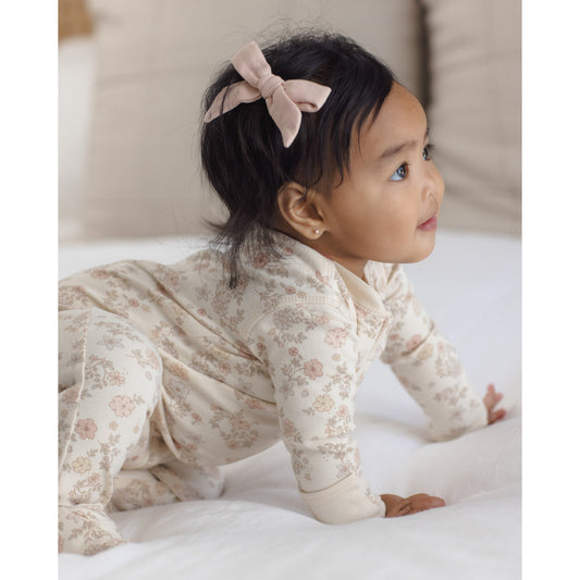 A baby crawling on a white bed wearing a cream-colored floral long-sleeve footed sleeper and a dusty pink bow in her hair.