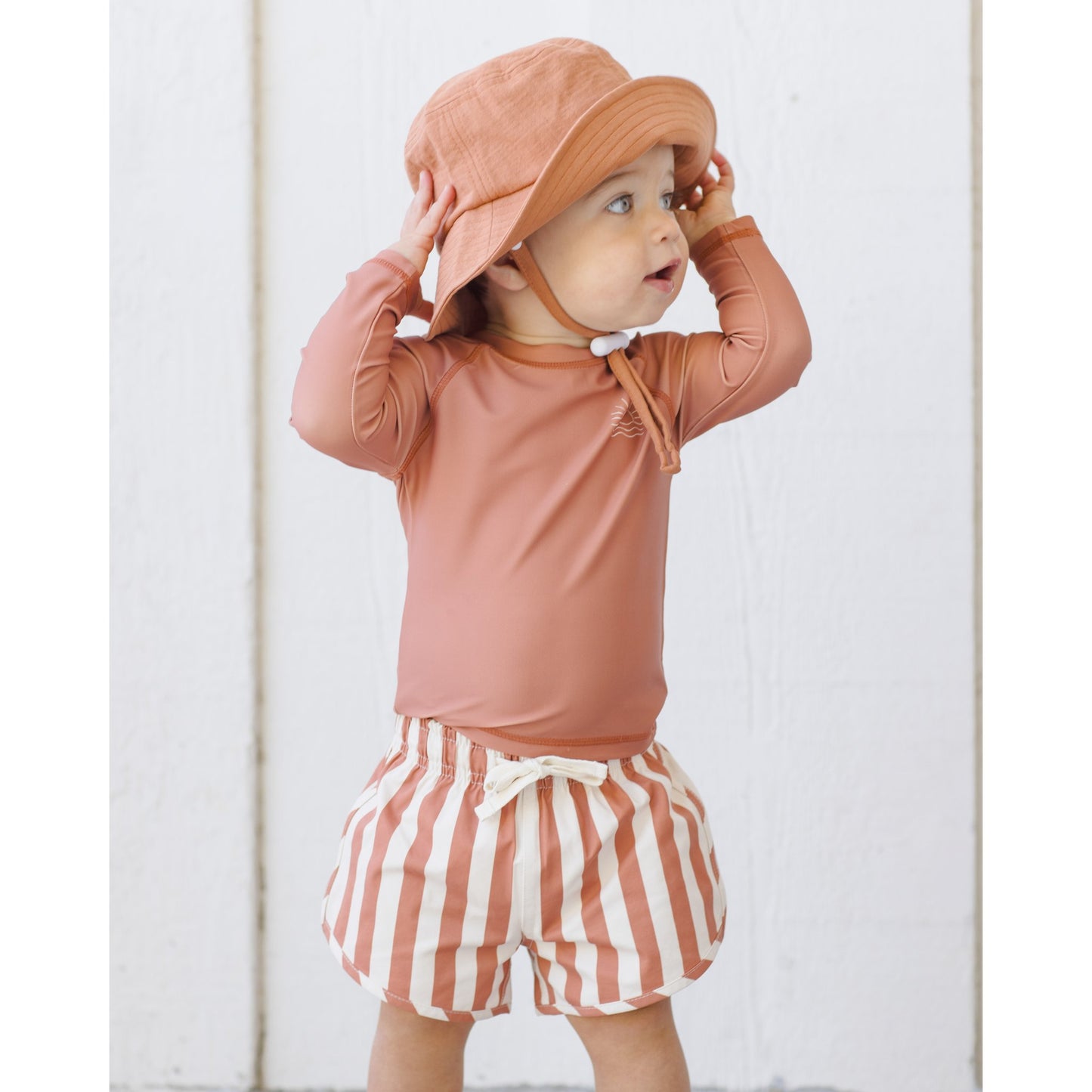 Child wearing a poppy long-sleeve shirt and striped shorts with a matching hat against a white background