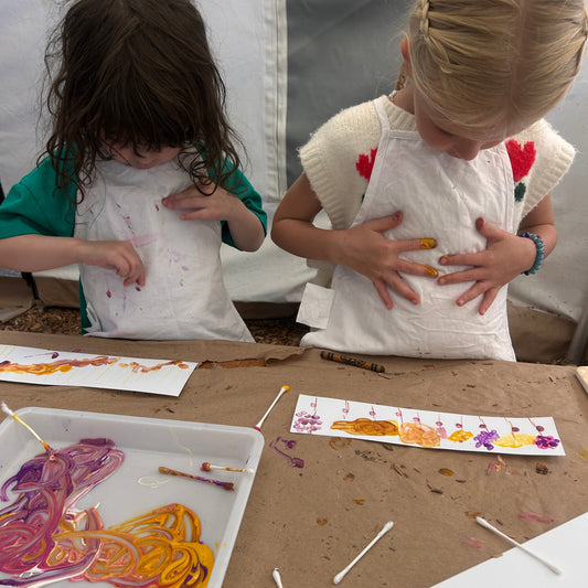 Two children wearing white aprons with colorful floral embroidery are painting on paper strips at a craft table. A white tray containing colorful string and paintbrushes sits in the foreground on a brown work surface.