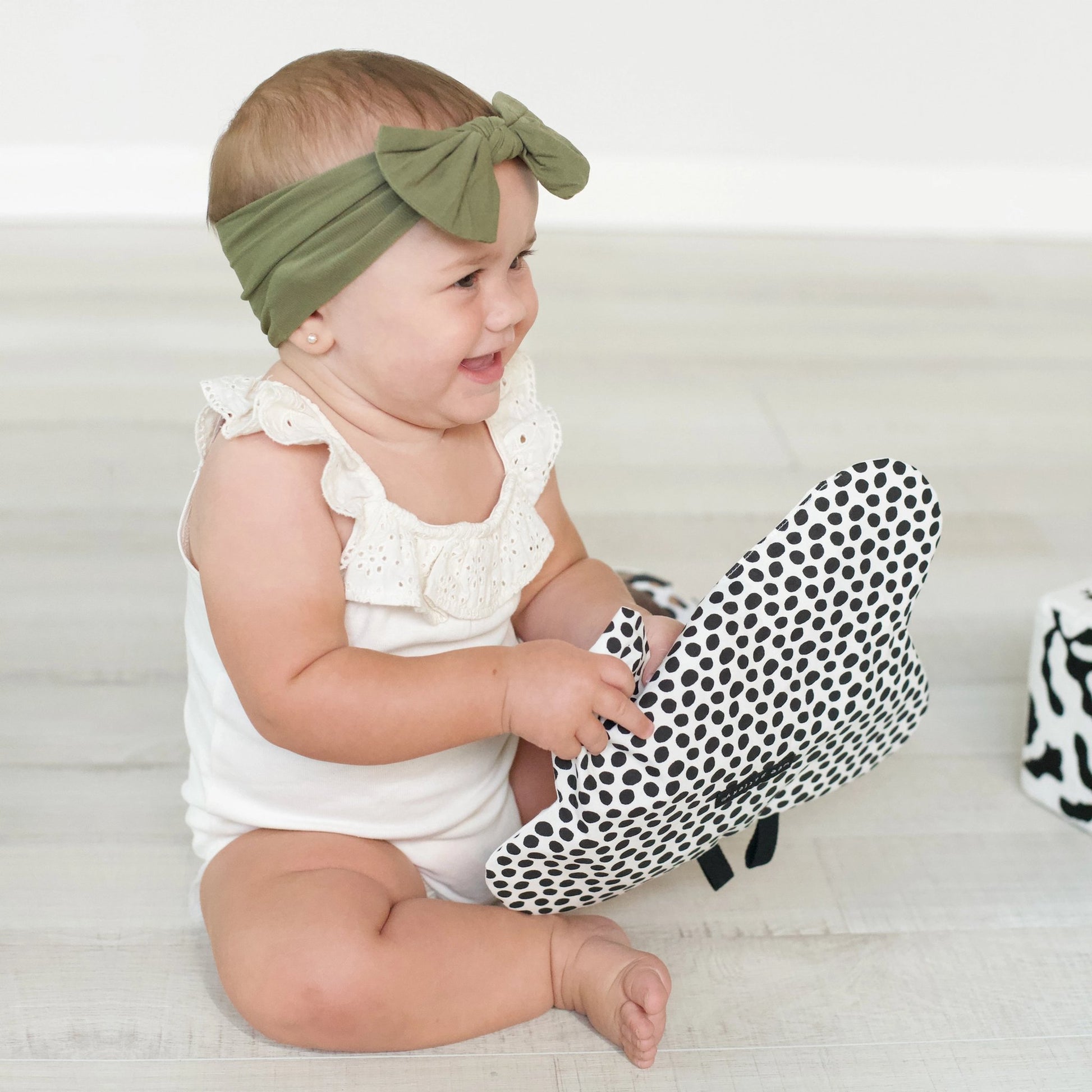 Baby sitting on a white surface wearing a green headband and holding a black and white polka dot egg crinkle toy